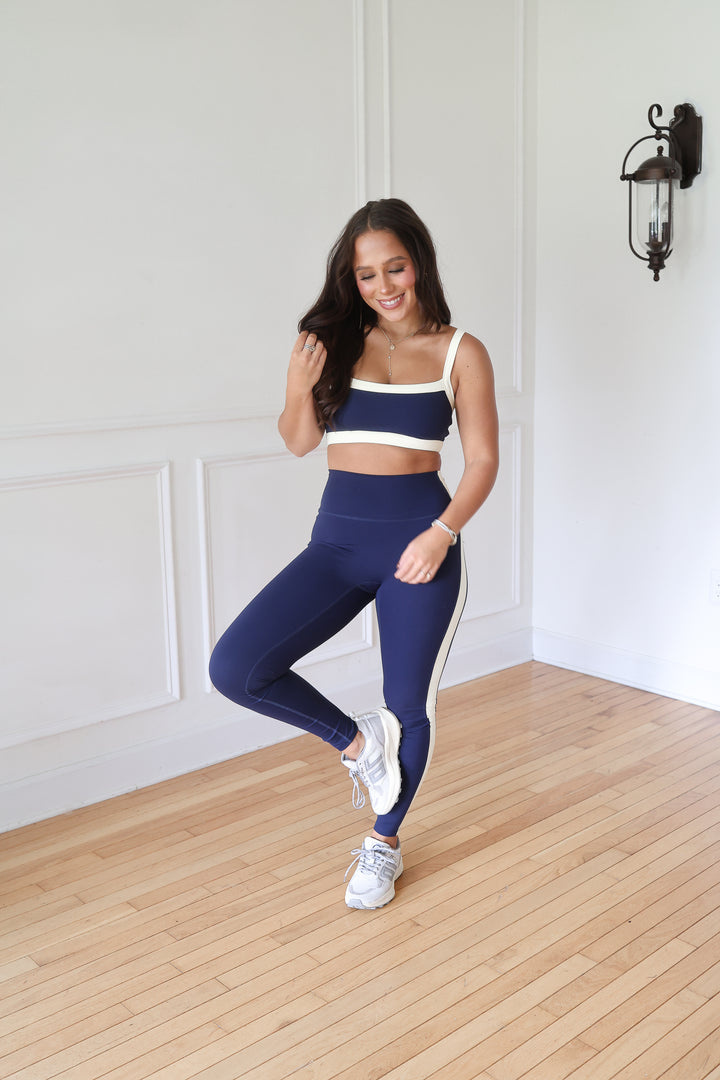 A woman modeling a matching navy blue activewear set consisting of a sports bra and high-waisted leggings with white side stripes