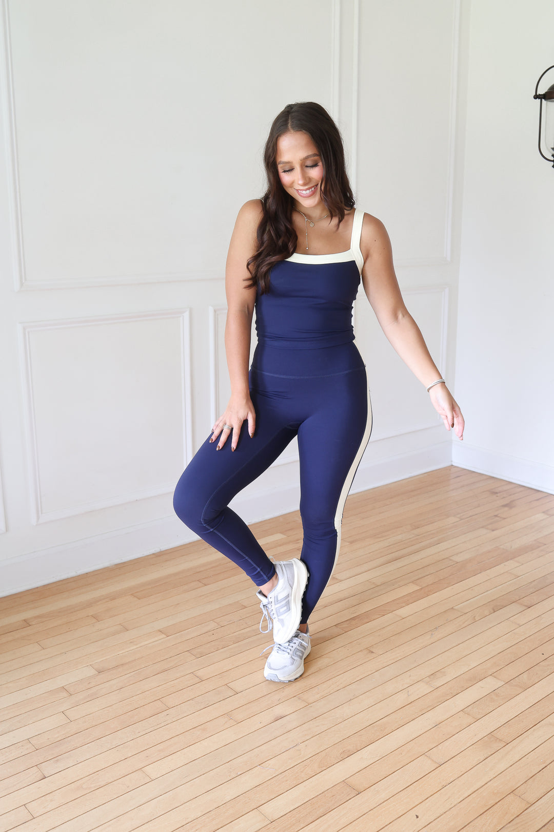 A woman modeling a navy blue ribbed activewear set consisting of a tank top and matching leggings with white contrast trim, paired with white sneakers.