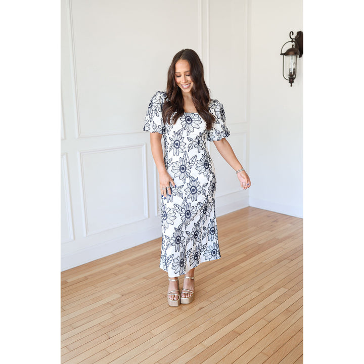 Woman wearing a floral dress standing in a room with wooden flooring and white walls.