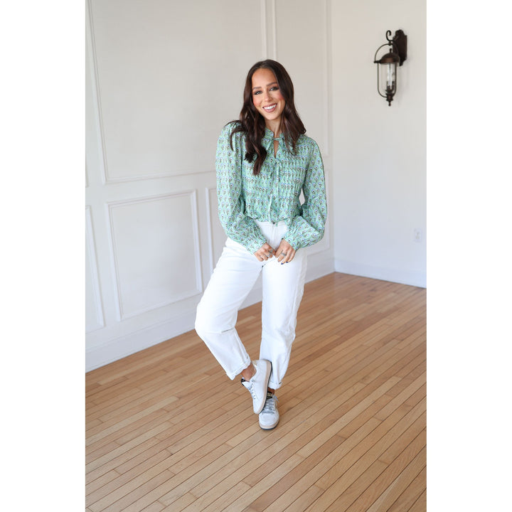 Woman in a green patterned blouse and white pants standing in a room with wooden flooring and white walls.