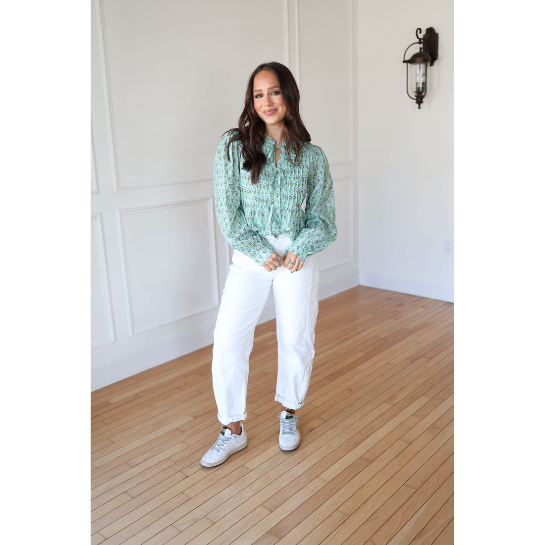 Woman wearing a green patterned blouse and white pants standing in a room with wooden flooring and white walls.