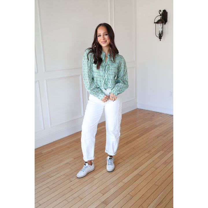 Woman wearing a green patterned blouse and white pants standing in a room with wooden flooring and white walls.