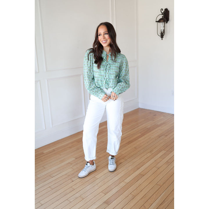 Woman wearing a green patterned shirt and white pants standing on a wooden floor.