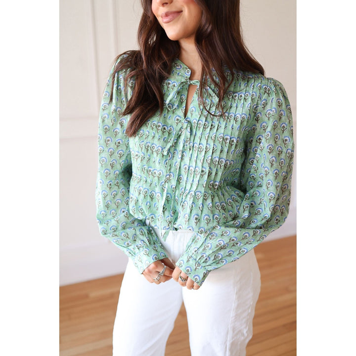 Woman wearing a green patterned blouse and white pants against a neutral background
