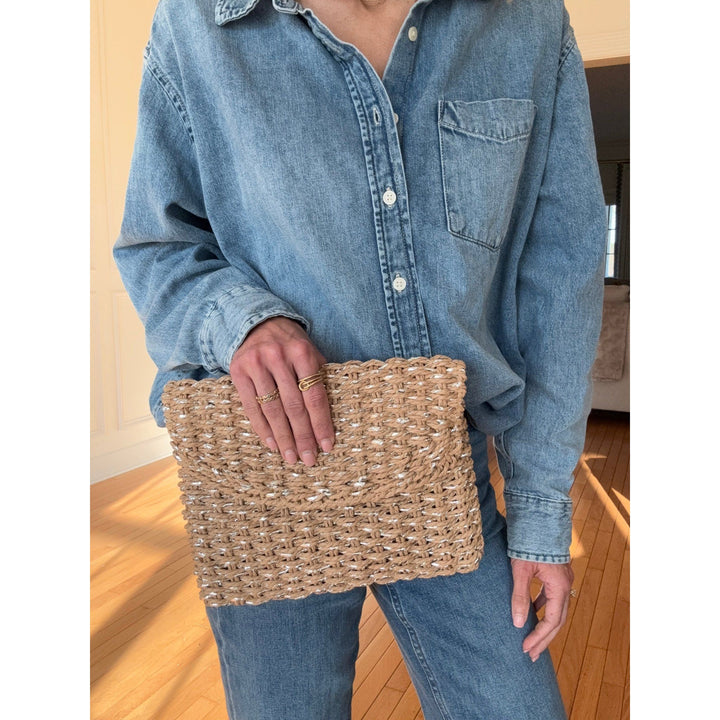 Person holding a woven clutch bag wearing a denim shirt and jeans indoors.
