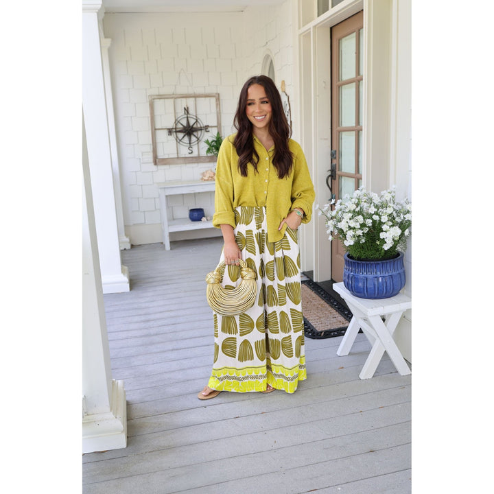 Woman in a yellow top and patterned pants standing on a porch with flowers and decor.