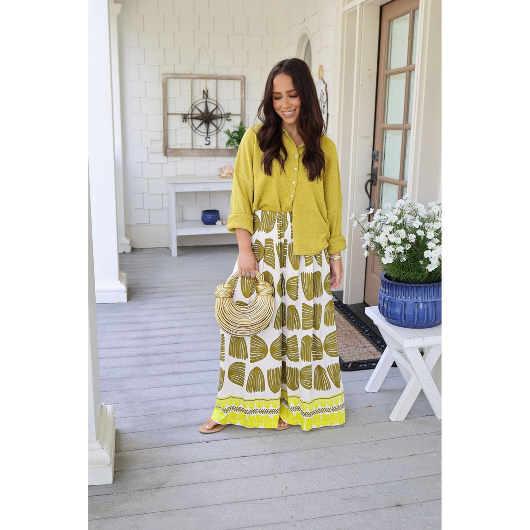 Woman in a yellow shirt and patterned skirt standing on a porch with a white wall and wooden door in the background.