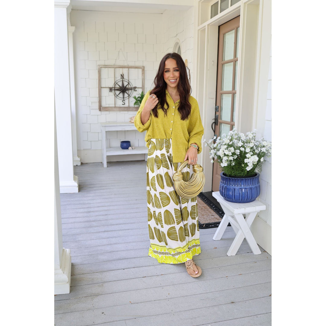 Woman in a yellow top and patterned skirt standing on a porch with white walls and wooden floor.