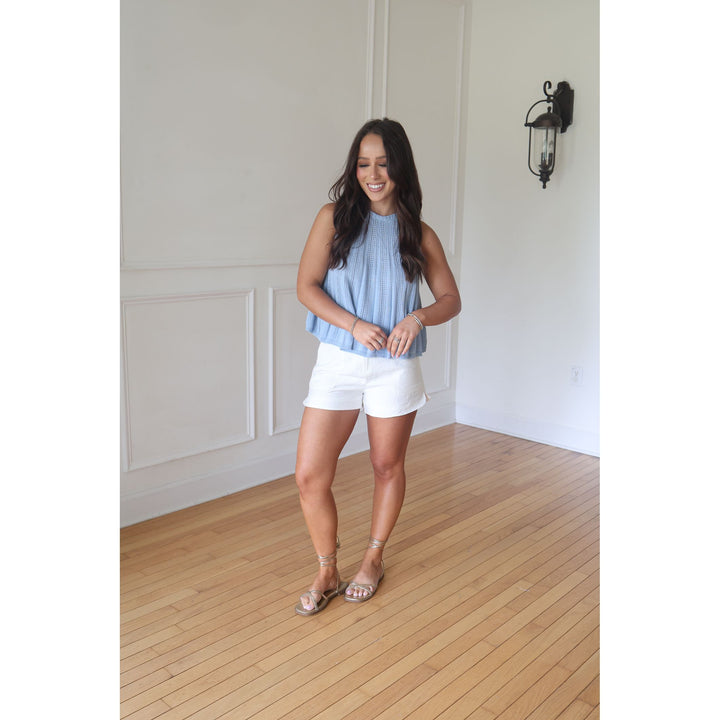 Woman wearing a light blue sleeveless top and white shorts standing in a room with wooden flooring and white walls.