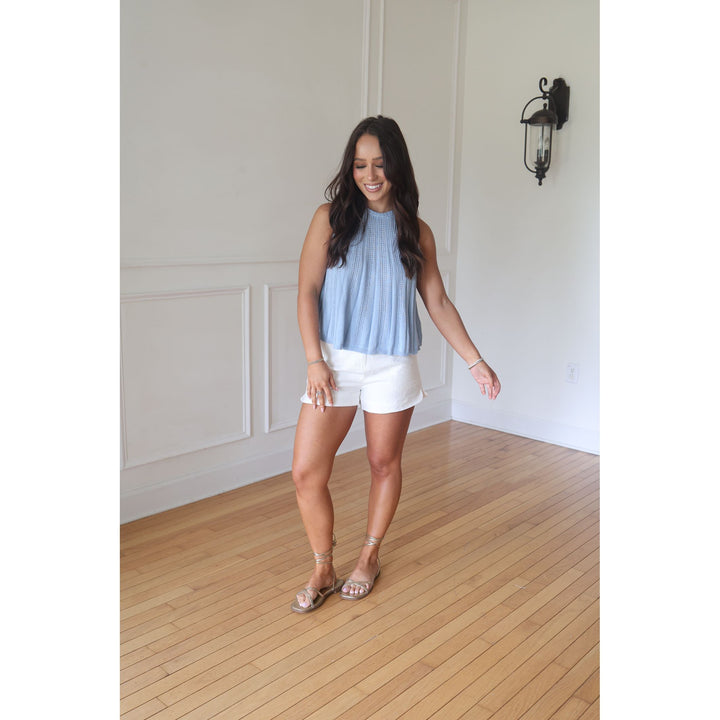 Woman in a light blue sleeveless top and white shorts standing in a room with wooden flooring and white walls.