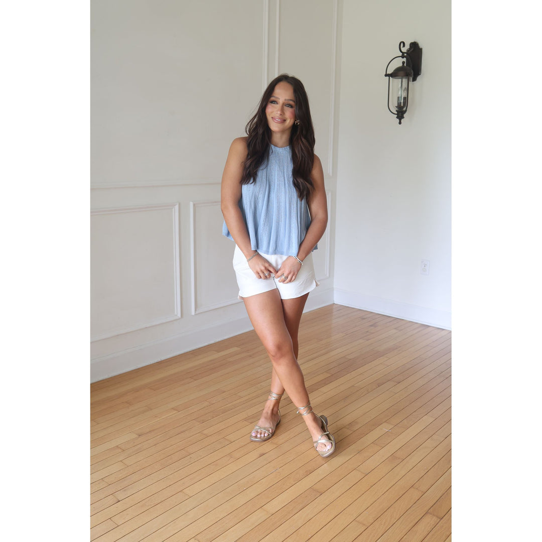 Woman in a light blue sleeveless top and white shorts standing in a room with wooden flooring and white walls.