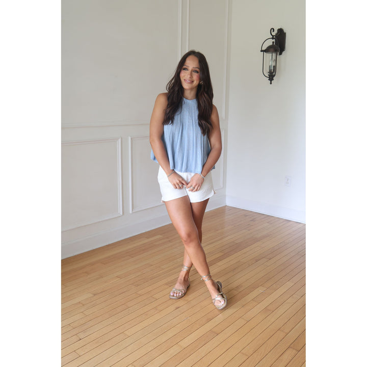 Woman in a light blue sleeveless top and white shorts standing in a room with wooden flooring and white walls.
