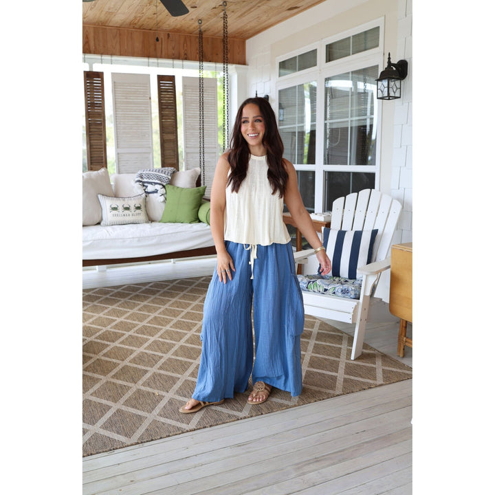 Woman standing on a porch wearing a white top and blue pants, with a house in the background.