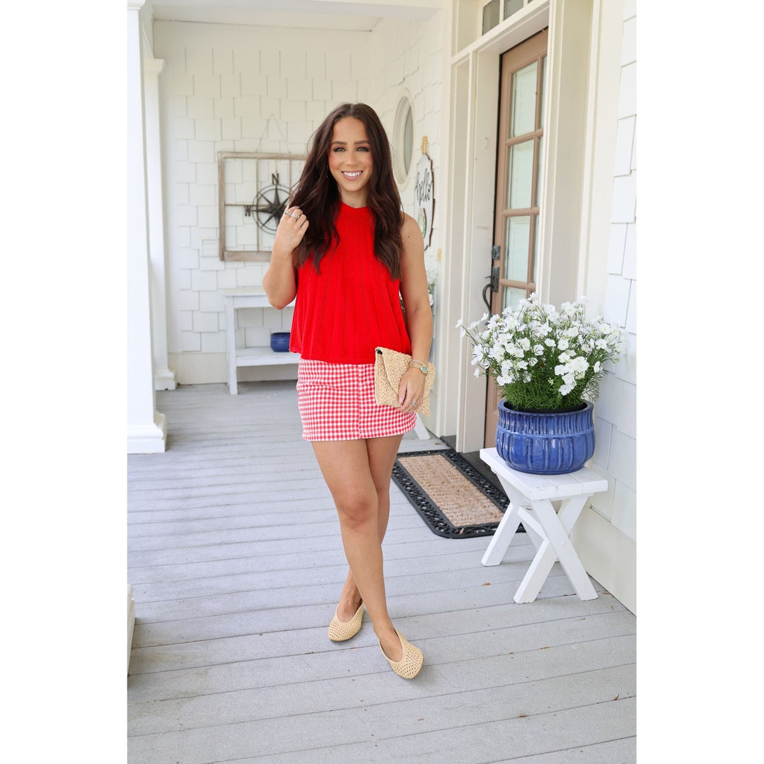 Woman in a red top and gingham skirt standing on a porch with a plant and door in the background.