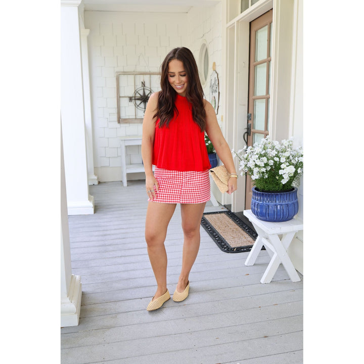 Woman in a red top and checkered skirt standing on a porch with plants and decor.